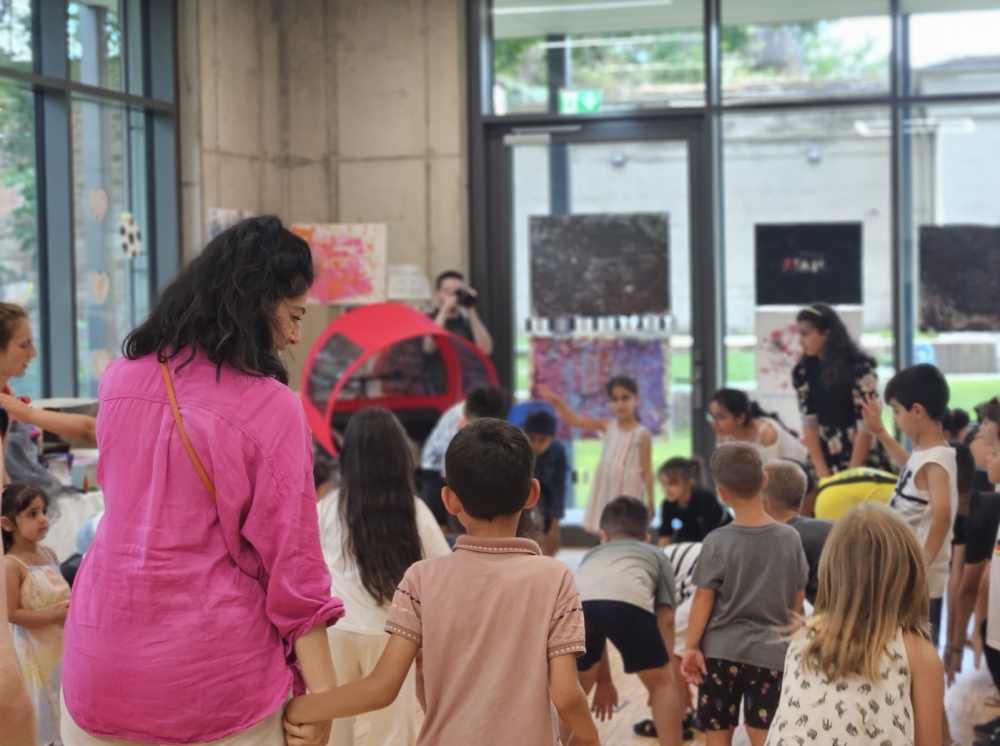 Abschlussfeier Sprachcamp Gruppe von Kindern und Erwachsenen stehen in Innenräumen der Bildungswerkstatt, rote Rikscha im Hintergrund zu sehen