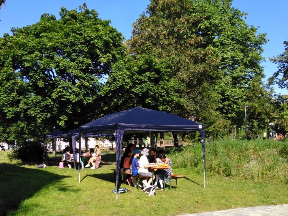 Sprachcamp 2024 Gruppe von Kindern und Erwachsenen sitzt am Tisch unter blauem Zelt auf grünem Außengelände