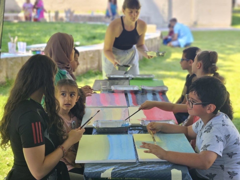 Kunstfreitag Gruppe von Kindern sitzt draußen an Tisch und malt auf Leinwänden, während eine junge Frau anleitet