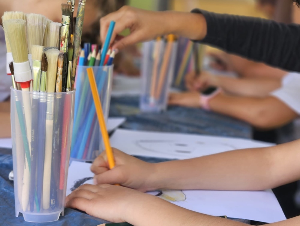 Kunstfreitag Becher mit Pinsel und Stiften stehen auf Tisch, Kinder arbeiten an Bildern