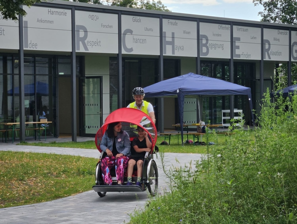 Sprachcamp 2024 Kind und Erwachsene sitzen in roter Rikscha, die von Mann gefahren wird, Gebäude Bildungswerkstatt und blaues Zelt im Hintergrund zu sehen
