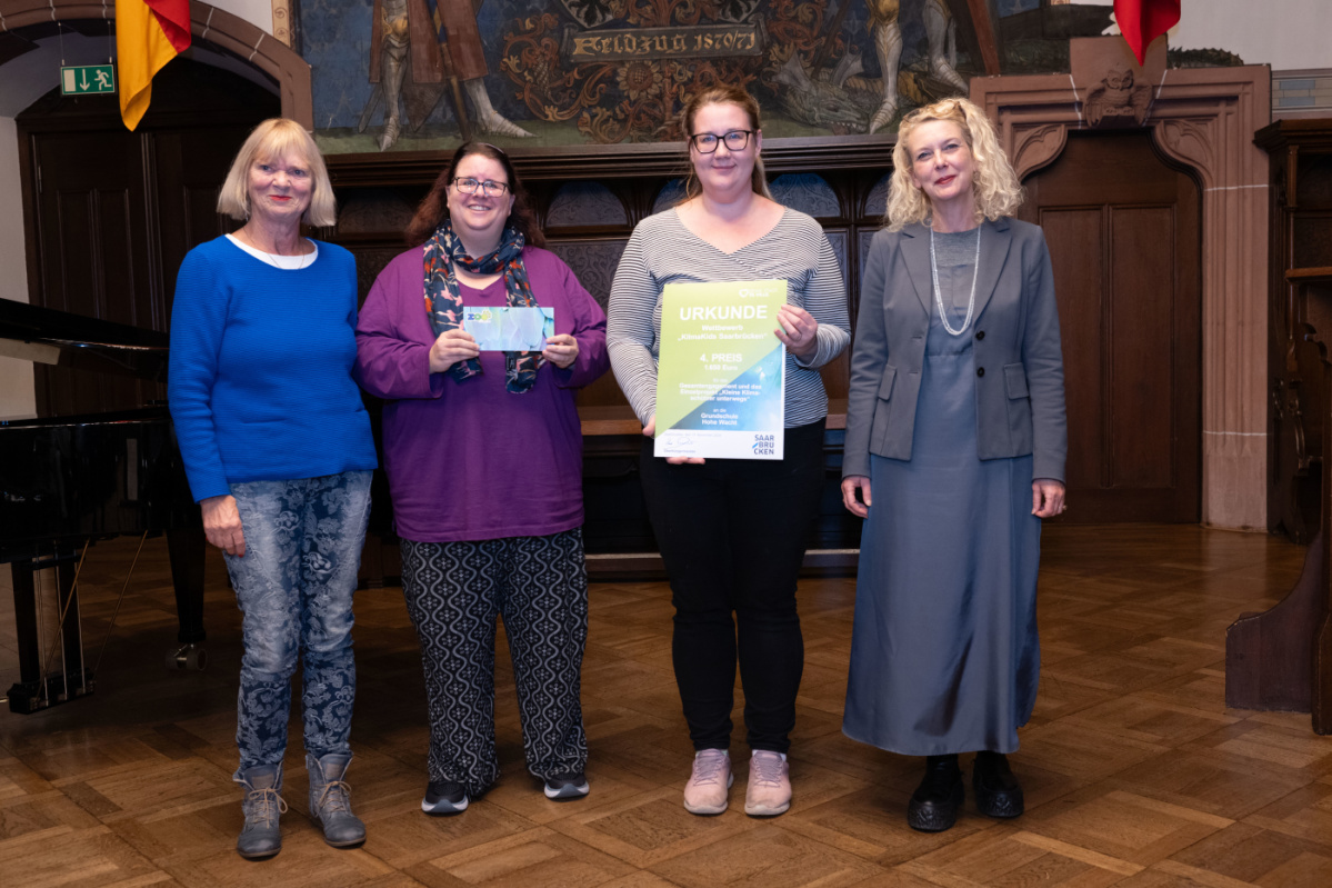 Platz 4 - Grundschule Hohe Wacht Preisübergabe des vierten Platzes an die Grundschule Hohe Wacht. Zu sehen sind zwei Betreuerinnen welche die Urkunde halten, sowie zwei Jurymitglieder.