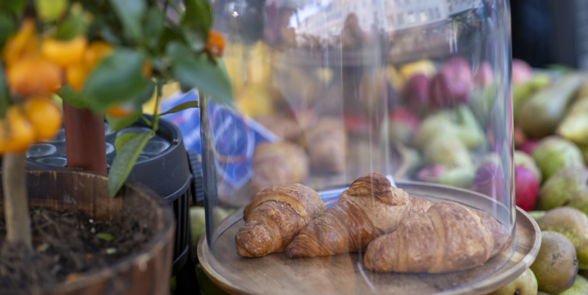 Zwei Croissants unter Glasglocke auf Marktstand, dahinter Früchte